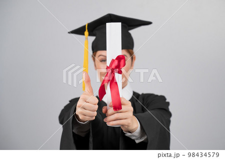 Happy woman in graduation gown holding out diploma and showing thumb up.  98434579