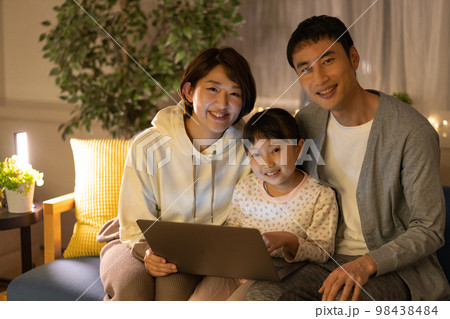夜　ソファに並んで座ってノートパソコンを見るパパとママと小学生の女の子 98438484
