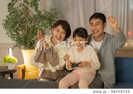 夜　ソファに並んで座ってテレビゲームをする小学生の女の子とパパとママ 98438539
