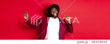 Christmas and people concept. Happy Black man celebrating New Year and dancing, wearing party outfit, red background 98439938
