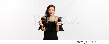 Fashion and online shopping concept. Happy young woman in black dress, showing credit card and mobile screen, standing over white background Fashion and online shopping concept. Happy young woman in black dress, showing credit card and mobile screen, standing over white background 98439950