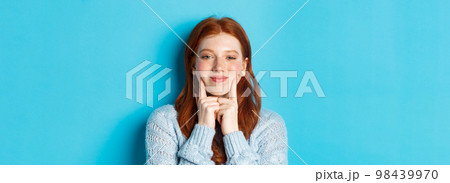 Close-up of cute and silly girl with red hair poking cheeks, showing dimples and smiling, standing against blue background 98439970