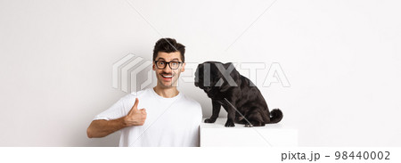 Image of happy and satisfied young man sitting near pug dog and showing thumbs-up, smiling and praising good product, white background Image of happy and satisfied young man sitting near pug dog and showing thumbs-up, smiling and praising good product, white background 98440002