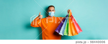 Covid-19, social distancing and lifestyle concept. Young man in face mask showing shopping bags and thumb-up, buying holiday gifts during pandemic Covid-19, social distancing and lifestyle concept. Young man in face mask showing shopping bags and thumb-up, buying holiday gifts during pandemic 98440006