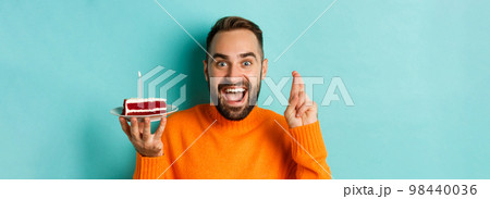 Close-up of happy adult man celebrating birthday, holding bday cake with candle and making wish, standing against turquoise background 98440036