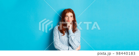 Pretty teenage girl with red hair thinking, looking upper left corner thoughtful, pondering something, standing over blue background 98440183