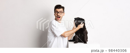 Happy young man showing you cute face of his pug. Dog owner loving his pet, standing over white background Happy young man showing you cute face of his pug. Dog owner loving his pet, standing over white background 98440306