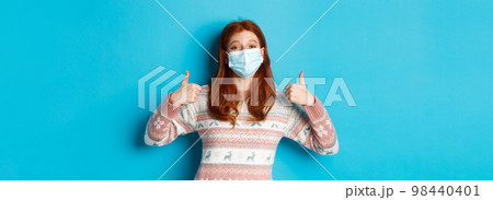 Winter, coronavirus and social distancing concept. Satisfied redhead girl in medical mask and sweater, showing thumbs up in approval, praise something good, blue background 98440401