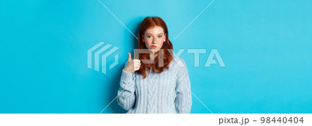 Sassy redhead girl in sweater, looking pleased and showing thumb up, like and agree, standing over blue background 98440404