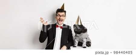 Happy young man and cute black dog wearing party cones, celebrating birthday, guy friendly saying hello and waving hand, standing over white background 98440405