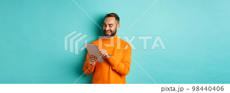 Handsome adult man working on digital tablet and smiling, shopping online, standing over light blue background Handsome adult man working on digital tablet and smiling, shopping online, standing over light blue background 98440406