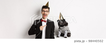 Happy dog owner and black pug wearing birthday party cones, man pointing at pug, standing over white background 98440502