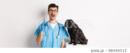 Handsome young doctor at vet clinic pointing finger up and looking amazed, standing near cute black pug dog, white background 98440513