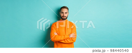 Confident young man looking determined, cross arms on chest, wearing orange winter sweater, standing against turquoise studio background Confident young man looking determined, cross arms on chest, wearing orange winter sweater, standing against turquoise studio background 98440540