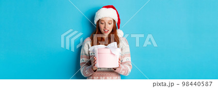 Winter and Christmas Eve concept. Touched and flattered redhead girl looking at box with xmas gift, smiling amazed, standing in santa hat against blue background 98440587