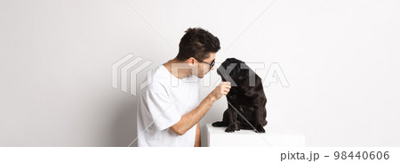 Handsome young hipster in glasses, pet his dog, looking at each other, standing over white background Handsome young hipster in glasses, pet his dog, looking at each other, standing over white background 98440606