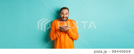 Handsome adult man celebrating birthday, blowing out candle on cake and making wish, standing against turquoise background 98440609