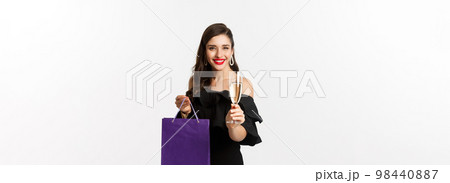 Beautiful and stylish brunette woman raising glass of champagne, celebrating christmas, holding shopping bag with presents, standing over white background 98440887