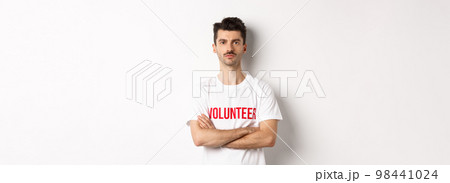 Serious young male volunteer in white t-shirt, holding arms crossed on chest, looking at camera, ready to help 98441024