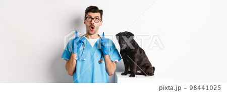 Handsome young doctor at vet clinic pointing fingers up and looking amazed, standing near cute black pug dog, white background Handsome young doctor at vet clinic pointing fingers up and looking amazed, standing near cute black pug dog, white background 98441025