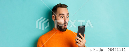 Close-up of caucasian man staring at phone screen with surprised face, wearing orange sweater, light blue background 98441112