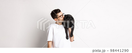 Image of handsome young man kissing his cute black pug, holding dog on shoulder, standing over white background Image of handsome young man kissing his cute black pug, holding dog on shoulder, standing over white background 98441290