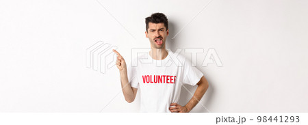 Disappointed volunteer showing bad promo offer, sticking tongue and grimacing disgusted, pointing finger left, wearing organization t-shirt, white background 98441293