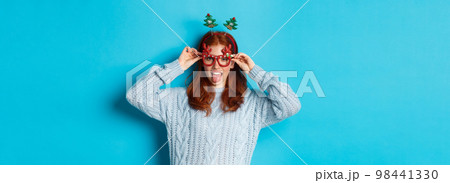 Winter holidays and Christmas sales concept. Beautiful redhead female model celebrating New Year, wearing funny party headband and glasses, smiling at camera 98441330
