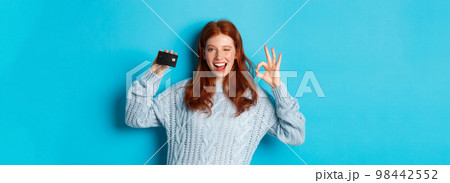 Happy redhead girl in sweater showing credit card and okay sign, recommending bank offer, standing over blue background Happy redhead girl in sweater showing credit card and okay sign, recommending bank offer, standing over blue background 98442552