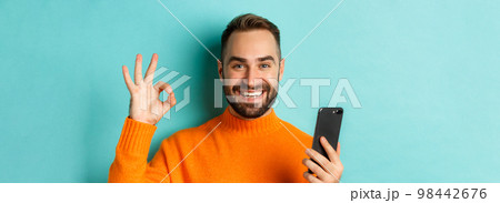 Close-up of bearded man holding smartphone and showing okay sign, recommending mobile app, standing satisfied over turquoise background 98442676