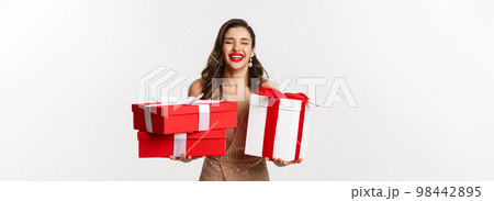 Holidays, celebration concept. Beautiful caucasian woman in elegant dress holding Christmas presents and smiling happy, standing over white background 98442895