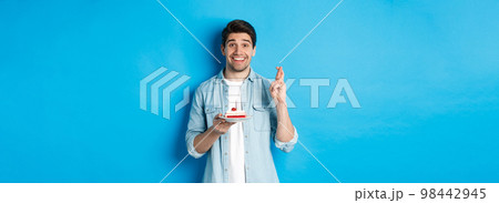 Hopeful man holding birthday cake and cross fingers, making wish, standing over blue background 98442945