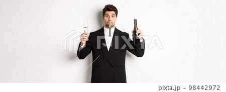 Concept of holidays, party and celebration. Portrait of handsome guy in black suit, raising bottle of champagne and glass, blowing a party whistle, having a birthday, standing over white background Concept of holidays, party and celebration. Portrait of handsome guy in black suit, raising bottle of champagne and glass, blowing a party whistle, having a birthday, standing over white background 98442972