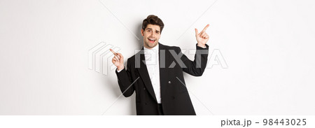 Handsome guy dancing and partying in black suit, pointing fingers sideways, showing two promo banners, standing against white background 98443025