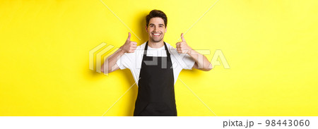 Smiling seller in black apron showing thumbs up, approve or like something, recommending cafe or store, yellow background Smiling seller in black apron showing thumbs up, approve or like something, recommending cafe or store, yellow background 98443060