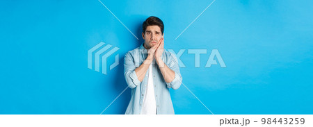 Portrait of man with toothache grimacing from pain and touching cheek, standing against blue background Portrait of man with toothache grimacing from pain and touching cheek, standing against blue background 98443259