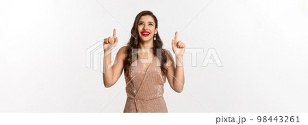 New Year, christmas and celebration concept. Happy elegant woman in party dress, laughing and looking up, pointing at top promo offer, white background New Year, christmas and celebration concept. Happy elegant woman in party dress, laughing and looking up, pointing at top promo offer, white background 98443261