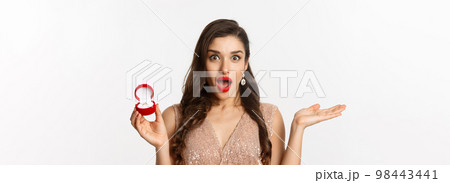 Close-up of attractive brunette woman with red lips, wearing dress, receiving engagement ring and looking surprise, standing over white background 98443441
