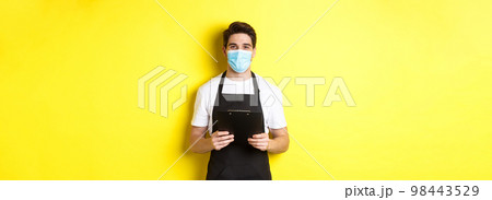 Concept of covid-19, small business and quarantine. Salesman in black apron and medical mask holding clipboard, working in shop, standing over yellow background 98443529