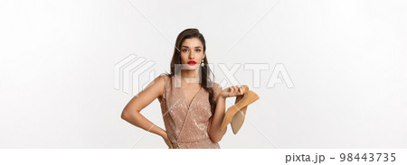 Party and celebration concept. Sassy and beautiful woman in luxury dress and red lipstick, holding pair of heels and looking unbothered, standing over white background Party and celebration concept. Sassy and beautiful woman in luxury dress and red lipstick, holding pair of heels and looking unbothered, standing over white background 98443735