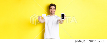 Handsome caucasian male model showing smartphone screen and credit card, concept of mobile banking and online shopping, yellow background 98443752