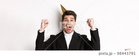 Close-up of happy good-looking man, wearing party hat and suit, raising hands up and rejoicing, celebrating new year, standing against white background Close-up of happy good-looking man, wearing party hat and suit, raising hands up and rejoicing, celebrating new year, standing against white background 98443791