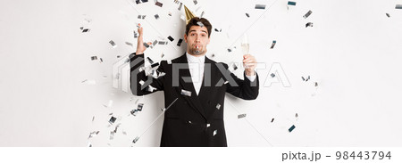 Handsome party guy in black suit having fun, celebrating new year, blowing whistle and drinking champagne while confetti falling, wishing happy holidays, standing against white background 98443794