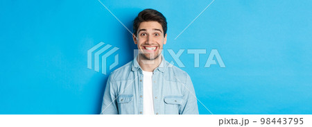 Close-up of smiling excited man with beard, looking amused at advertisement, standing against blue background 98443795