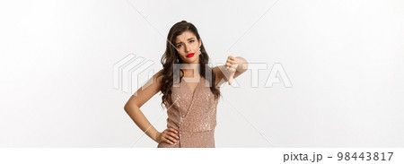 Skeptical young woman in elegant dress looking disappointed, showing thumbs-down, dislike christmas party, standing over white background Skeptical young woman in elegant dress looking disappointed, showing thumbs-down, dislike christmas party, standing over white background 98443817