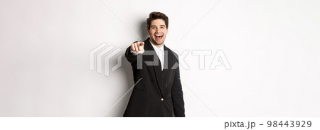 Concept of new year party, celebration and lifestyle. Portrait of handsome stylish man in black suit, smiling and pointing finger at camera, standing over white background 98443929