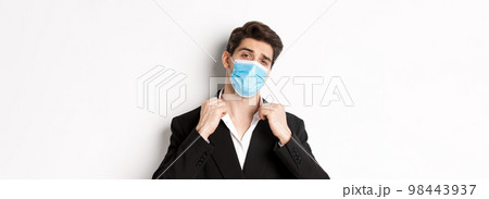 Concept of covid-19, business and social distancing. Image of confident handsome man in trendy suit and medical mask, looking sassy, standing against white background Concept of covid-19, business and social distancing. Image of confident handsome man in trendy suit and medical mask, looking sassy, standing against white background 98443937