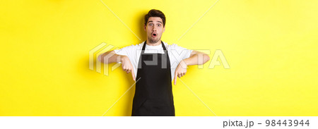 Surprised seller in black apron pointing fingers down, looking amazed at camera, standing over yellow background 98443944