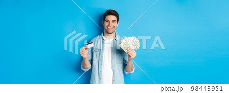 Young smiling man showing cash dollars and credit card, standing over blue background 98443951