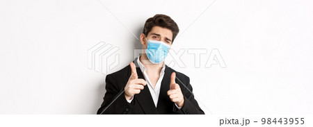 Concept of covid-19, business and social distancing. Close-up of sassy man in trendy suit and medical mask, pointing fingers at camera, standing against white background 98443955